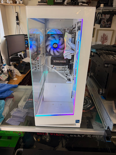 A white custom PC tower with a clear side panel, showcasing internal components and RGB lighting, set on a glass desk in a cluttered room.