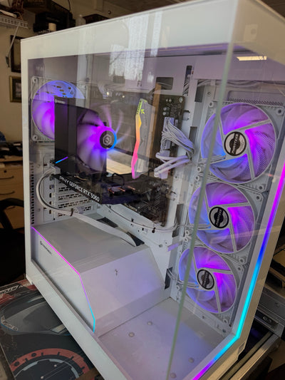 A white custom PC tower with a clear side panel, showing internal components including a GPU, RAM, and multiple RGB-lit fans. The case is photographed on a desk in a home or office environment.