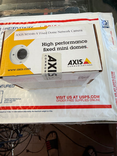 A boxed AXIS M3046-V Fixed Dome Network Camera sitting on top of a USPS shipping box. The product box is white with yellow and red accents and features the AXIS Communications branding.