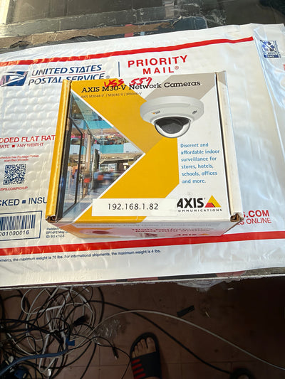 A 4Axis M30V network camera retail box with a white dome security camera pictured on the front, placed on top of a USPS Priority Mail bag. A person's foot in a black-and-red slipper is visible at the bottom edge.