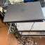 A closed gray Dell laptop sits on a metal stand in a cluttered electronics workspace with cables, keyboards, and a blue cloth visible.