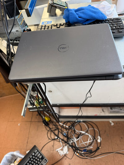 A closed gray Dell laptop sits on a metal stand in a cluttered electronics workspace with cables, keyboards, and a blue cloth visible.